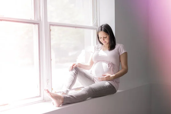 Young beautiful pregnant woman sitting on windowsill at home — Stock Photo, Image