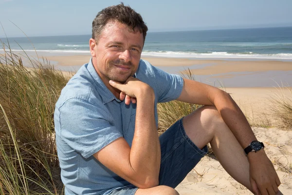Uomo bello e sicuro di sé sorridente e seduto in spiaggia — Foto Stock