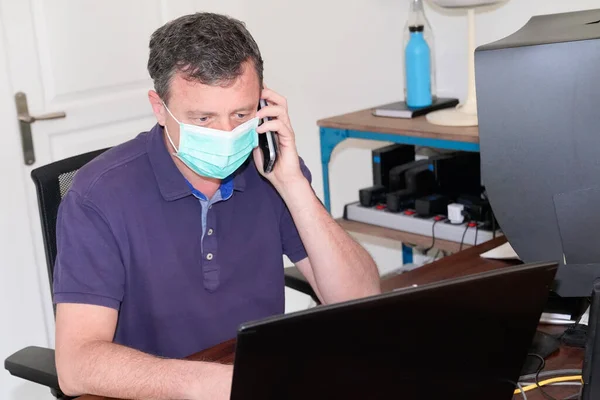 handsome man in protective face mask is talking on phone working computer from home office during Coronavirus COVID-19 virus epidemic quarantine disease