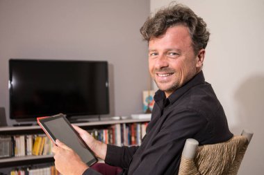 Evde oturan çekici yakışıklı adam elinde tablet bilgisayarla oturuyor.