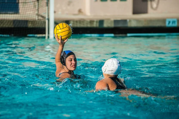 Female water polo athlete holding ball above water and away from defense while looking to score.