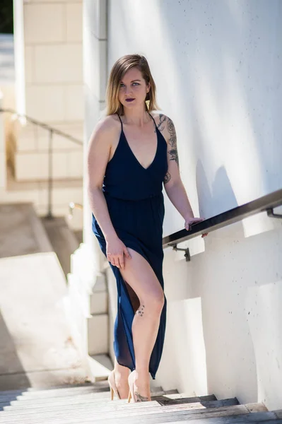 Alluring young woman in sheer blue dress ascending stairs with sexy leg revealed.