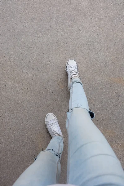 Τα πόδια της νεαρής γυναίκας της μόδας σε μπλε τζιν και λευκά αθλητικά παπούτσια στο πάτωμα — Φωτογραφία Αρχείου