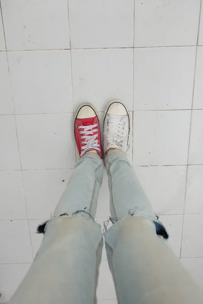 Τα πόδια της νεαρής γυναίκας της μόδας σε μπλε τζιν και λευκό, κόκκινα αθλητικά παπούτσια στο πάτωμα — Φωτογραφία Αρχείου