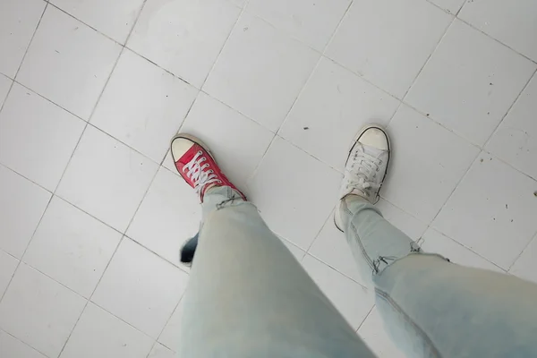 Gambe da donna di moda giovane in blue jeans e bianco, scarpe da ginnastica rosse sul pavimento — Foto Stock