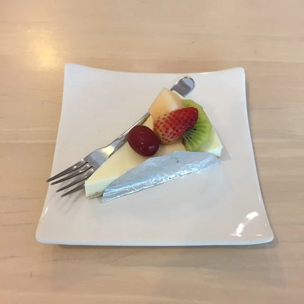 Fetta di torta di formaggio condita con composta di fragole, kiwi, cereali e melone sul piatto sul tavolo di legno con forchetta da dessert — Foto Stock