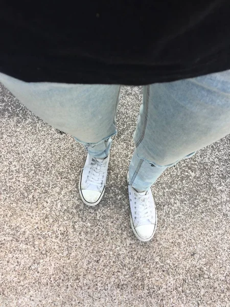 蓝色牛仔裤中的女腿和地板上的白色运动鞋 — 图库照片
