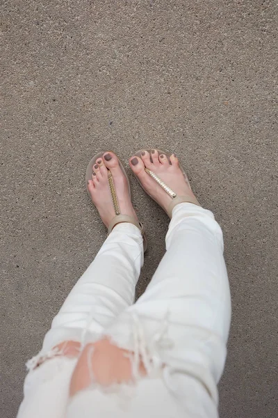 Selfie Feet Wearing Gold Sandals and White Jeans on Ground Background