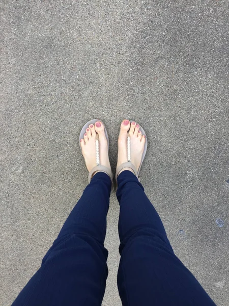 Close-up van de voeten met bruin nagel in gouden sandalen en blauwe broek op vrouwelijke voeten op de grond achtergrond — Stockfoto