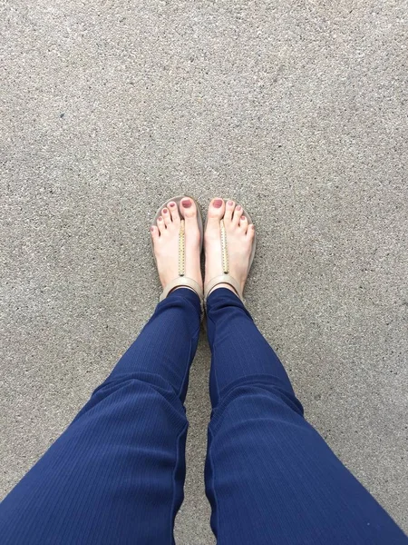 Close Up Feet with Brown Nail in Gold Sandals and Blue Pants on Female Feet on Ground Background