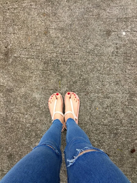 Close up of Bare Feet with Red Nail in Sandals and Blue Jeans Woman on the Concrete Floor Background — стоковое фото