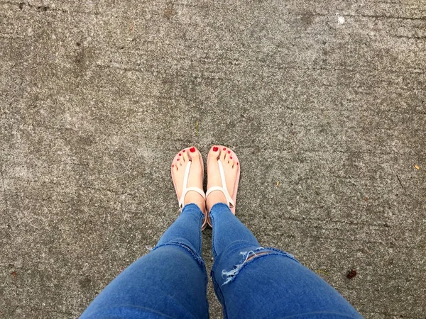 Close-up de pés descalços com prego vermelho em sandálias e jeans azuis Mulher no chão de concreto Fundo — Fotografia de Stock