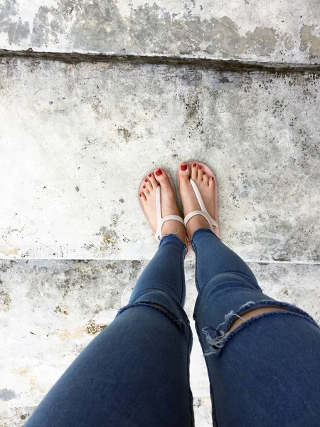 Primo piano di piedi nudi con chiodo rosso in sandali e Blue Jeans donna sullo sfondo del pavimento di cemento — Foto Stock