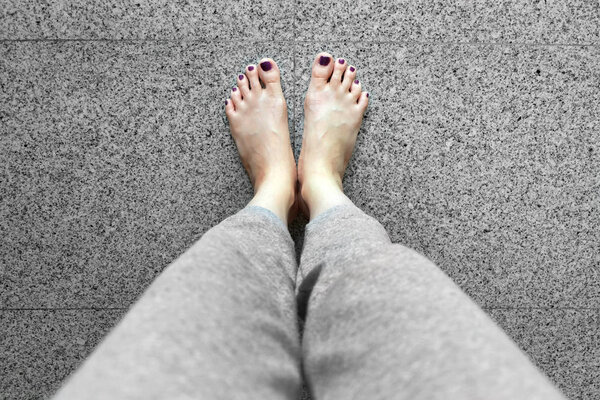 Босиком с педикюром для ногтей. Ноги женщины крупным планом носят серые брюки, стоящие на фоне серой плитки
