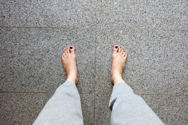 Босиком с педикюром для ногтей. Ноги женщины крупным планом носят серые брюки, стоящие на фоне серой плитки — стоковое фото