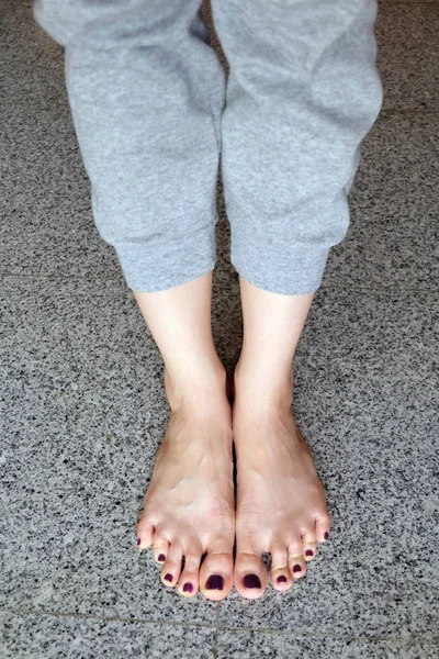 Босиком с педикюром для ногтей. Ноги женщины крупным планом носят серые брюки, стоящие на фоне серой плитки — стоковое фото
