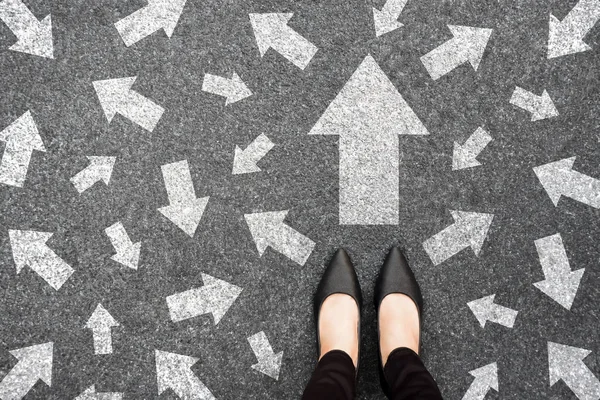 Feet and arrows on road background from above. Businesswoman standing on pathway with drawn white many direction arrows choice. Top view of A businesswoman black shoes. Motivation and growth concept.