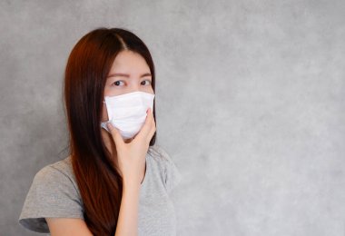 Gri arka planda koruma maskesi takan Asyalı bir kadın. İnsanlar cerrahi maske takıyor çünkü pm2.5 hava kirliliği, virüs gribi, koronavirüs ve COVID-19 ateşi var. Sağlık hizmetleri kavramı.