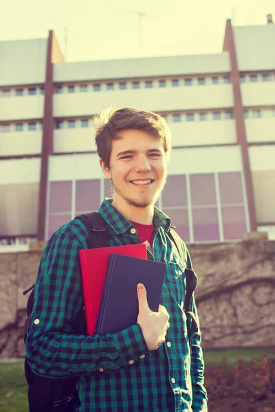 Un jeune étudiant souriant tenant un livre et un sac sur un fond universitaire — Photo