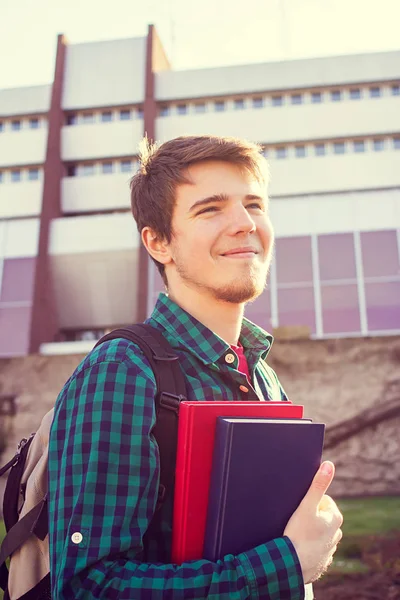 Un jeune étudiant souriant tenant un livre et un sac sur un fond universitaire — Photo