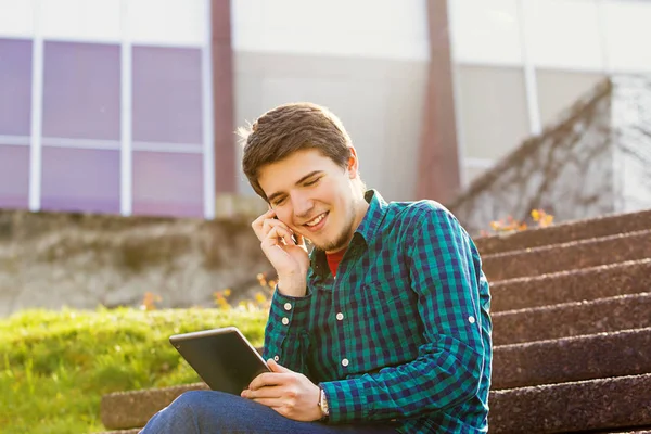 Un jeune homme souriant enregistre sur une tablette et parle sur un téléphone portable dans une ville par un escalier — Photo