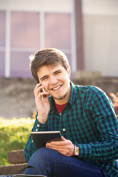 Un jeune homme souriant enregistre sur une tablette et parle sur un téléphone portable dans une ville par un escalier — Photo
