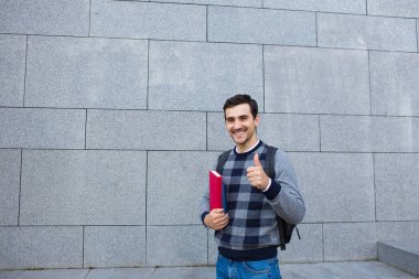 University.Smiling genç öğrenci manshowing başparmak yukarı ve bir kitap ve bir çanta Üniversitesi arka plan üzerinde tutan