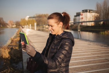 Nehri yakınında şehir köprüsü üzerinde otururken tablet kullanarak gülümseyen genç kadın, tablete, taping öğrenci.