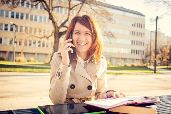 Smiling young woman student talking on mobile phone using tablet in campus university