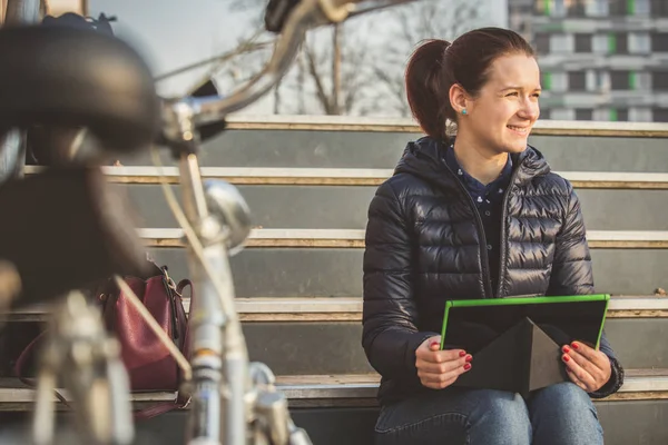 Uśmiechający się młoda kobieta, student, taping na tablecie przy użyciu tabletu w parku miejskim, siedząc na schodach w pobliżu rower — Zdjęcie stockowe