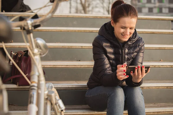 Smiling young woman, student  using mobile phone in a city park sitting on stairs near a bicycle
