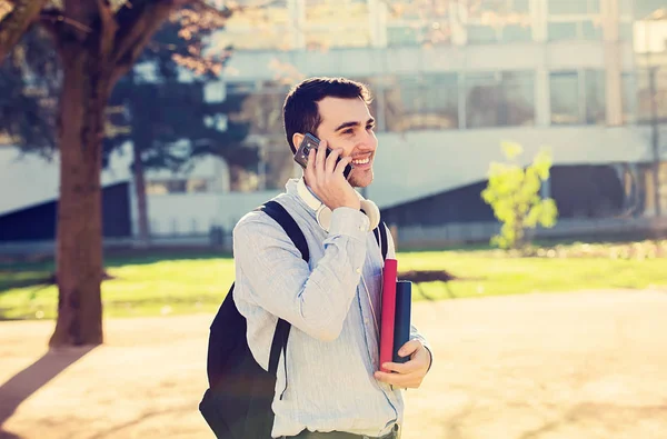 Souriant jeune étudiant hommeparler sur téléphone mobile tenant un livre et un sac sur un fond universitaire — Photo