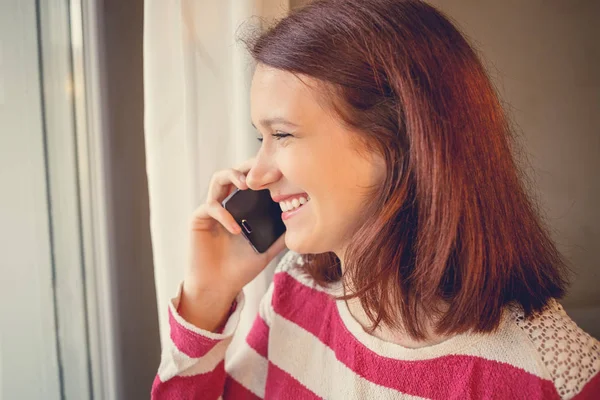 Smiling young woman student talking on mobile phone near a window home