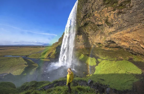 Mulher sob a cachoeira Seljalandsfoss, Islândia. Dia ensolarado na Islândia com arco-íris — Fotografia de Stock