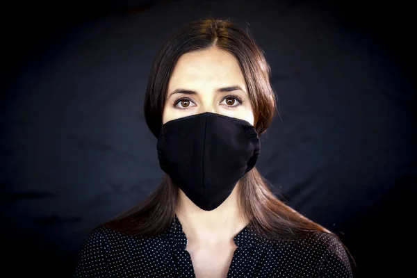 Young woman wearing a protective black leather face mask during the Coronavirus disease COVID-19 outbreak epidemic. Close up studio portrait on a black background with a multiple use protection mask on the face.
