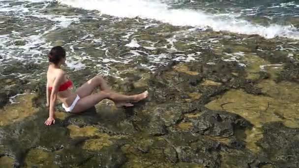 Молода красива дівчина в купальнику посміхається, сидить на скелі на узбережжі — стокове відео
