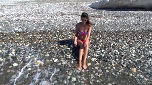 Chica sentada en la playa y mojándose los pies debido a una ola — Vídeos de Stock