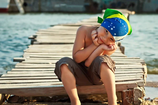 Chłopiec w chustka siedzi na drewniany most w piasku słoneczny letni dzień — Zdjęcie stockowe
