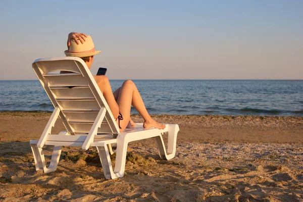 Vista trasera chica usando teléfono inteligente acostado en la chaise longue de la playa . — Foto de Stock