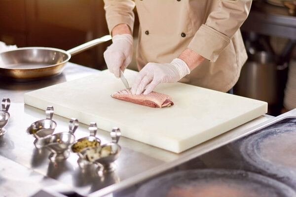 Chef cutting the meat on board. Male chef in gloves processing meat with knife at professional kitchen. Preparing delicious poultry fillet.
