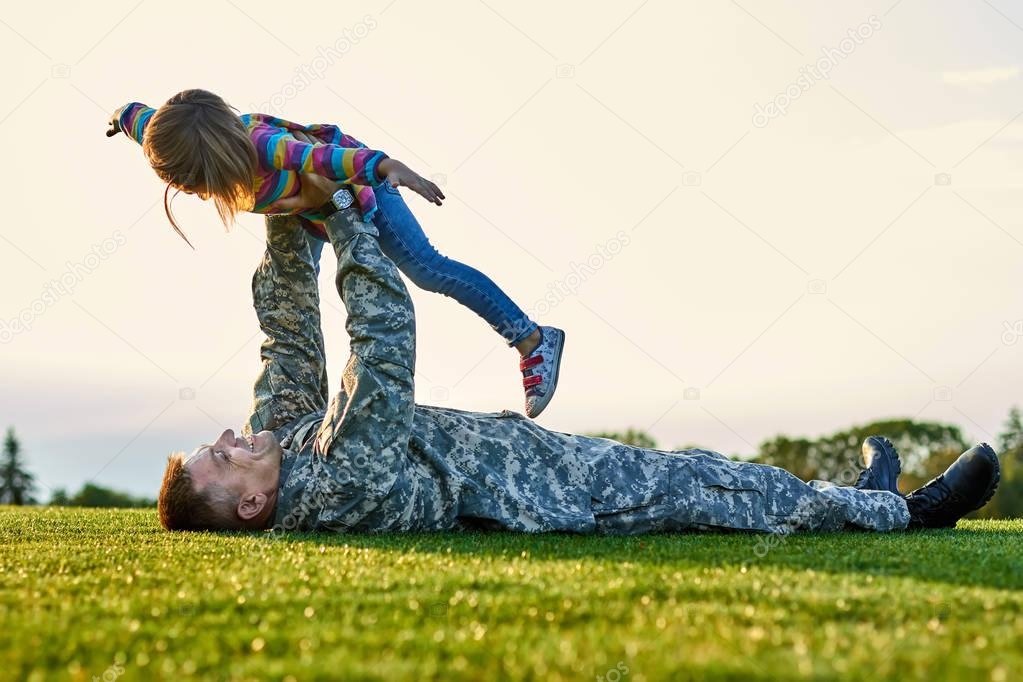 Soldier lying on the grass and playing with little girl.