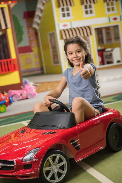 Happy little smiling girl sitting on toy car indoor. — ストック写真