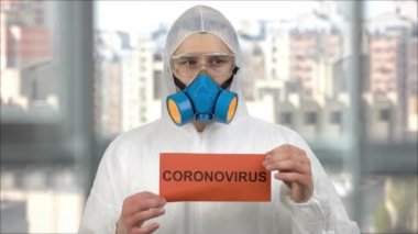 Koruyucu giysiler içinde portait od adam ve Coronavirus kelime kağıt tutan solunum maskesi.