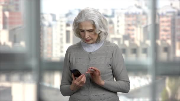 Пожилая женщина использует смартфон в помещении с городским пейзажем . — стоковое видео