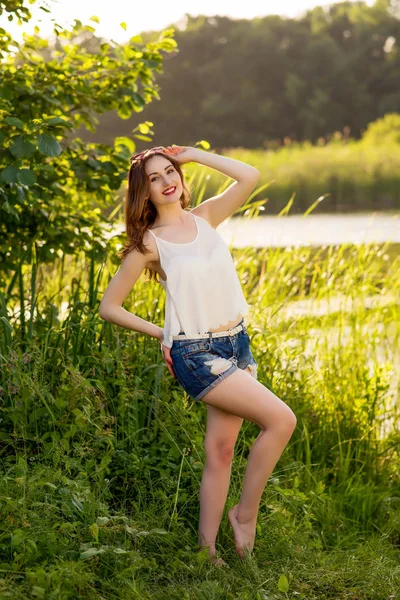 Menina sorridente bonita em uma camisa branca e calções de ganga em pé perto do rio — Fotografia de Stock