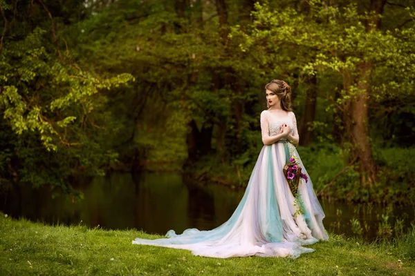 Fantastic bride with flowers in a long beautiful wedding dress stands near river