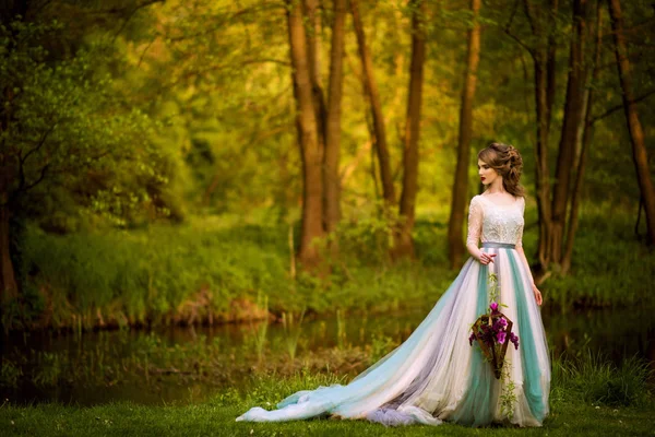 Fantastische bruid met bloemen in een lange mooie trouwjurk staat in de buurt van rivier — Stockfoto