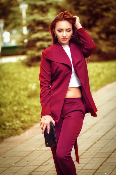 Belle jeune femme à la tête rouge portant une veste rouge, un sac à main en cuir, marchant dans la rue. Photo de mode — Photo