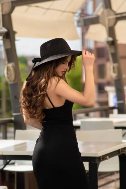 Lüks restoranda poz veren şapkalı ve siyah elbiseli muhteşem bir kadın. Güzel genç bayan. Kıvırcık saçlı moda kız.