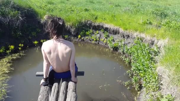 Joven con rastas se sienta en la orilla del lago y disfruta de la naturaleza — Vídeo de stock